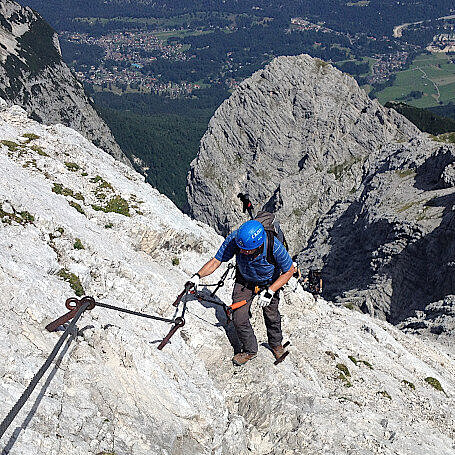 The author with helmet climbing up a via ferrata. Seen from above with Bavarian landscape below in the background.