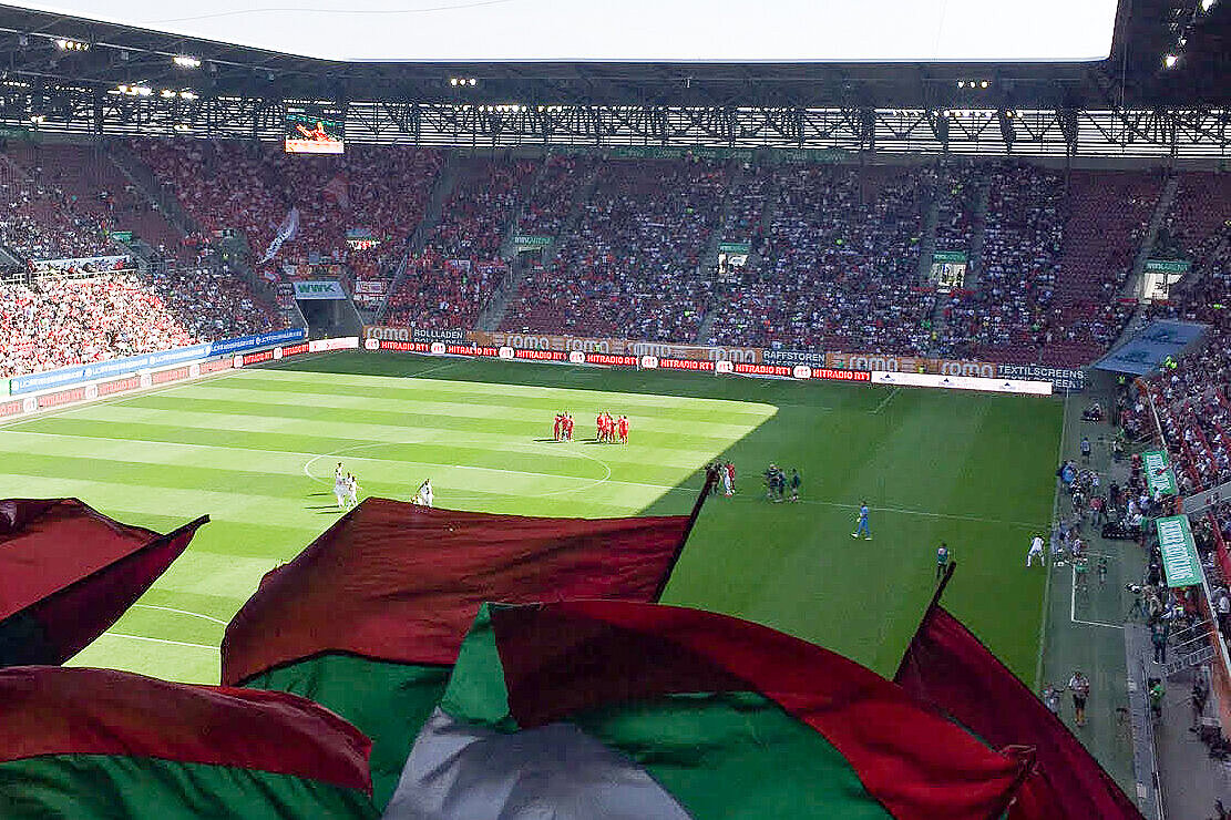 View from the tribune on the playground with players from FC Augsburg, flags of the team in the foreground.