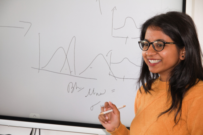 Researcher standing in front of a whiteboard with a pen in her hand and smiling at the audience. 