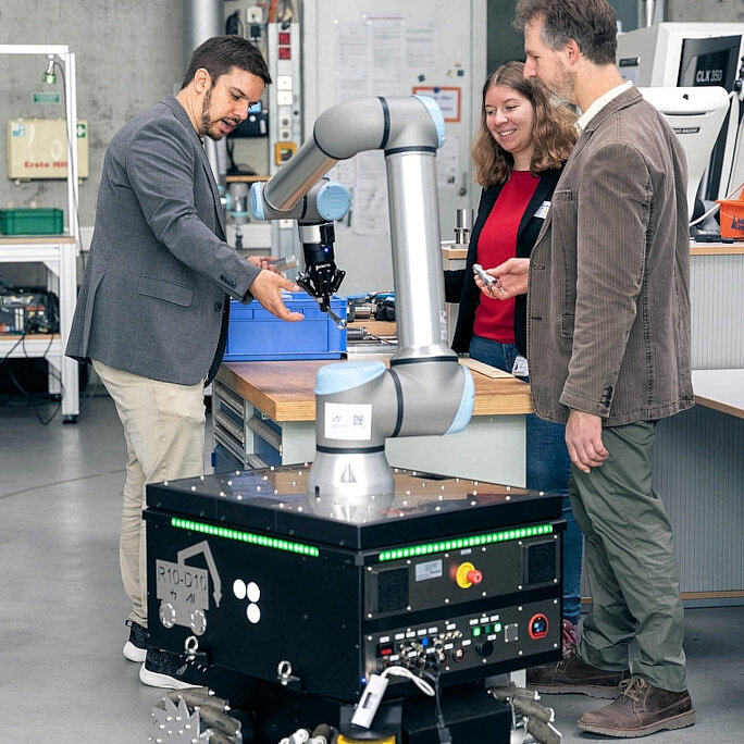 Three researchers testing a robot with wheels and a robot arm made for use of ai in production.