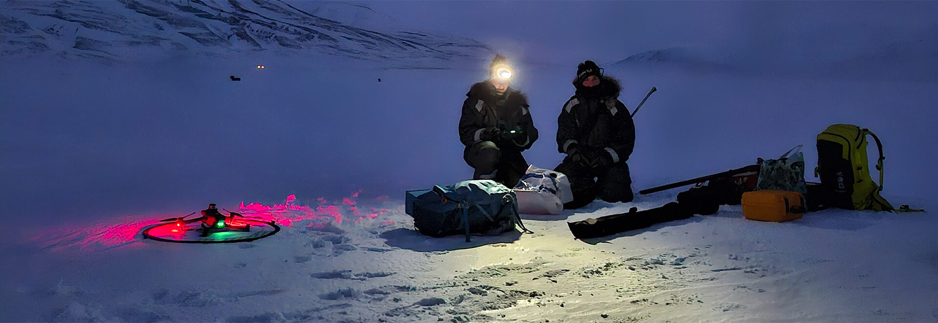 Two researchers are sitting in the middle of a snow-filled landscape without light. A pink and green lit drone lies next to them. Around the researchers are bags and equipment. One of the researchers has a flashlight on their head and is controlling the drone. 