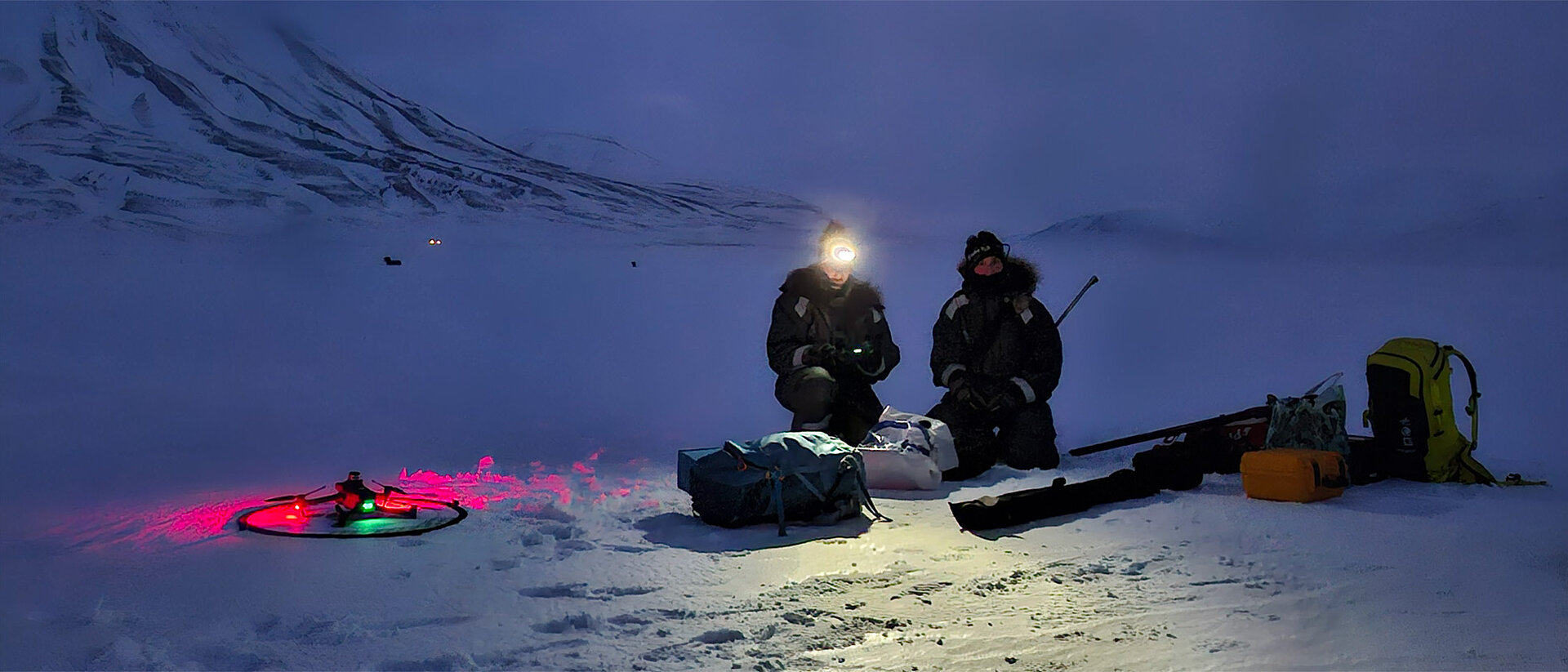 A researcher is shining light onto a small plane-shaped drone in the middle of snow-covered landscape. The drone is gray with neon green patterns.