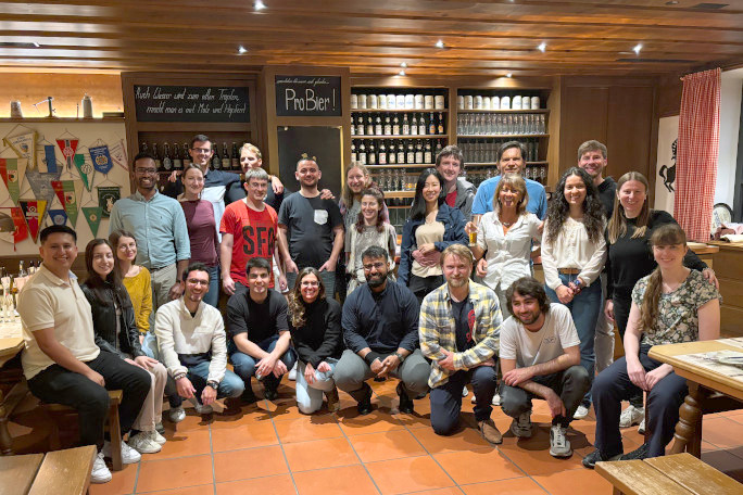 A big group of international researchers pose for a picture in a pub, on the wall behind them are beer bottles and mugs. A sign above the head of Filip Simonovski who stands in the center says "ProBier". 