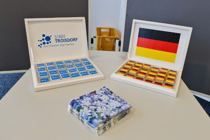 On a white table are two open boxes full of small nicely wrapped chocolate pieces. The box on the left says in German "Troisdorf City - a family matter" in blue writing, the chocolate wrappers also in blue. Box on the right has German flag printed on the box cover and on the chocolate wrappers. In front of the two boxes are napkins with floral patterns.