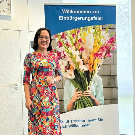 Chau Bui dressed in colorful Vietnamese traditional costume "ao dai" posing in front of a standee saying in German "Welcome to the Naturalization Ceremony", "Troisdorf City warmly welcomes you". On the standee is a picture of a bouquet of colorful flowers.