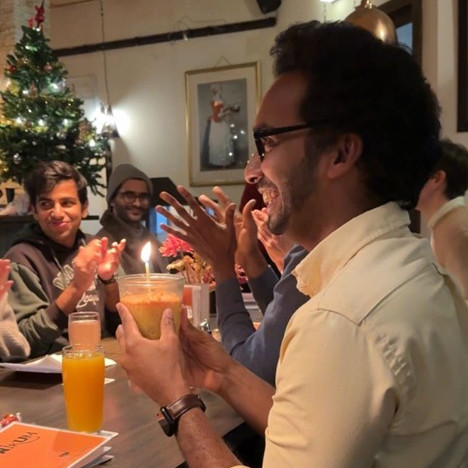 A photo of Daksitha smiling holding a small birthday cake with a lit candle sitting at the Stammtisch while people around him are clapping. In the background there is a well-lit and decorated Christmas tree and a picture on the wall.