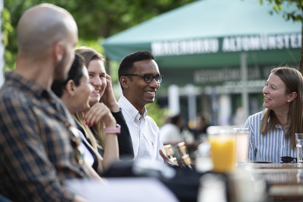 Daksitha sits between 4 friends at an outdoor table. He is captured smiling talking to his friends.