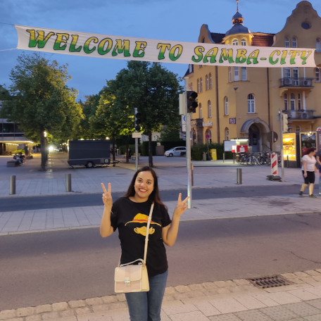 Snehanjali making V peace signs on both hands in front of a banner saying"Welcome to Samba-City" 