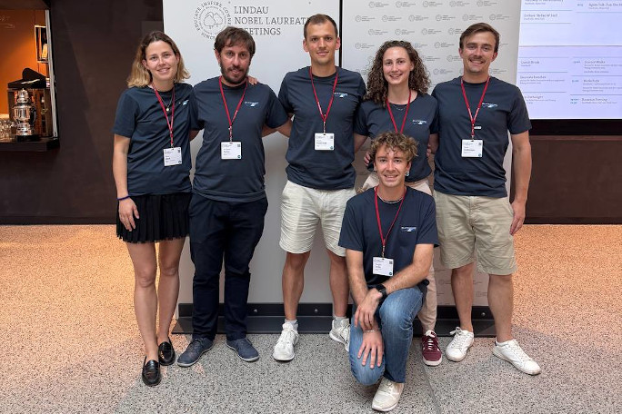 A group of 6 researchers in team SolTech smilingly pose in front of the Lindau Nobel Laureate Meetings backdrop, Morgan Le Dú kneels one knee down, posing in the first row.