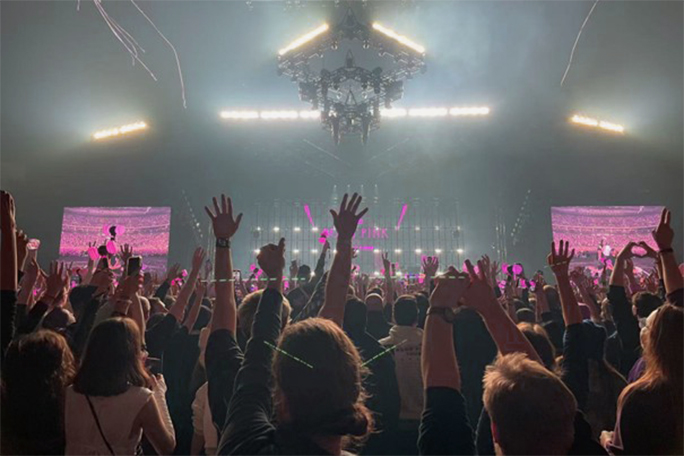 A concert scene with a lot of people putting their hands up. A big stage with bright lights and two side screens in pink is in the far background.