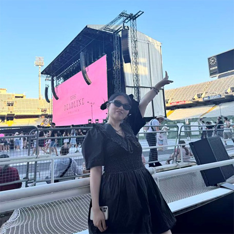 Yixiao Zhang joyously makes a V-sign with her left hand in the sky while posing in front of the concert stage with a big pink screen, leaning against the arm rails of the seating part in the stadium. Behind her are some people sitting facing the other way.