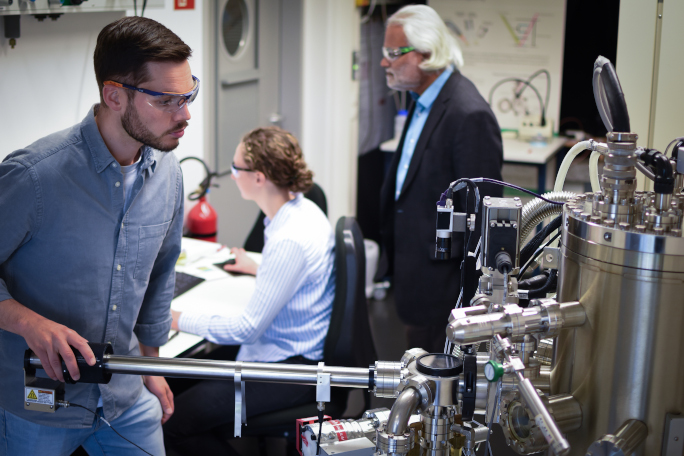 Young researcher using a machine to research solar energy conversion, a professor and student work in the background.