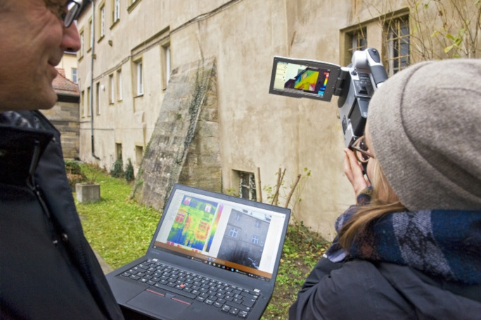 Two people standing outside, examining the surroundings with a thermal camera and a laptop. These are the usual tools in digital heritage conservation.
