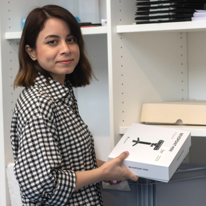 The young researcher standing in front of a shelf, with a box in her hands. a camera is pictured on it.