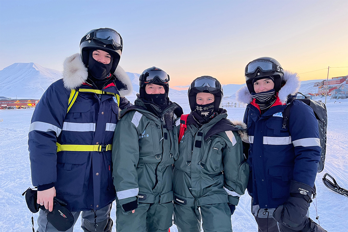 A group of 4 researchers in full gear smile at the camera. They all stand in a snow-covered landscape ful of light, in the background are small colorful houses..