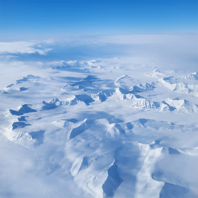 The aerial view of Svalbard on a bright day: white snow-covered mountains ridges are clear to see with only some clouds.
